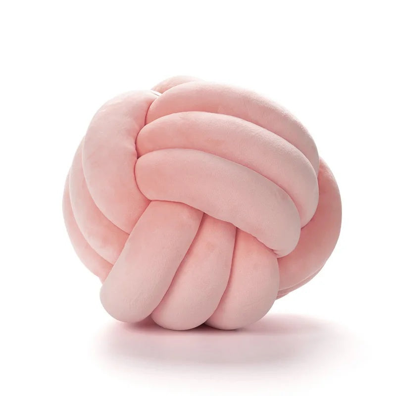Un grand coussin décoratif pour lit de bébé en forme de nœud, de couleur rose poudré, en tissu velours doux, posé sur un tapis rayé noir et blanc à franges devant un berceau blanc dans une chambre d'enfant de style scandinave