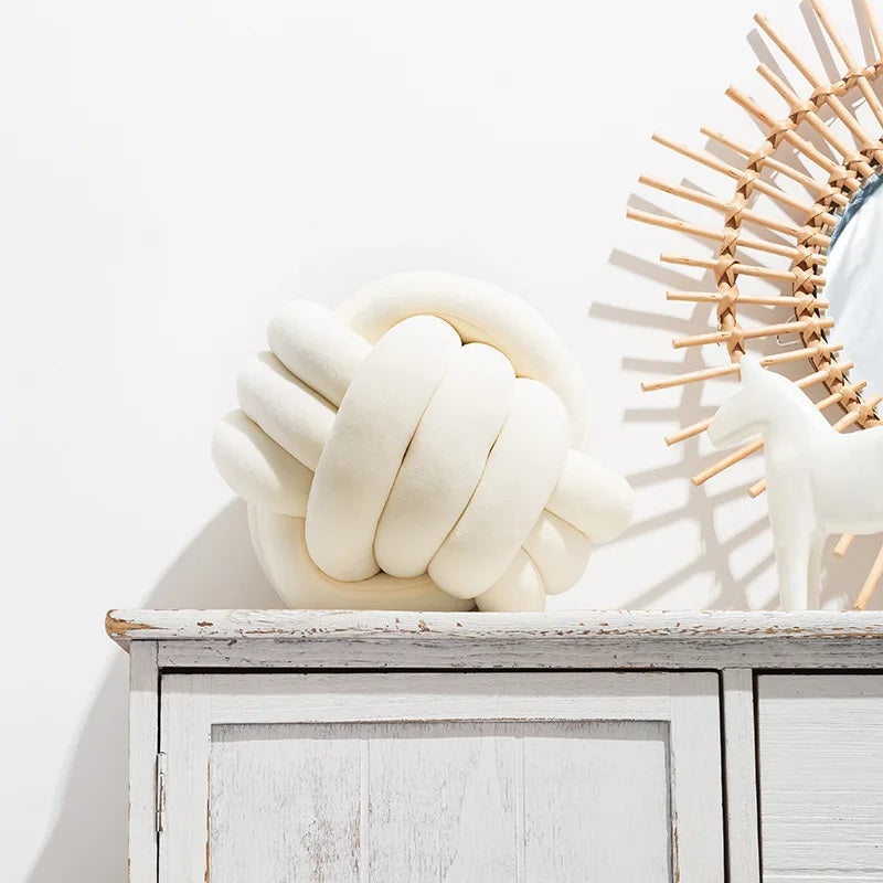 Un grand coussin décoratif pour lit de bébé en forme de nœud tressé, de couleur crème ou blanc cassé, posé sur un tapis rayé noir et blanc, devant un berceau blanc dans une chambre d'enfant de style scandinave.
