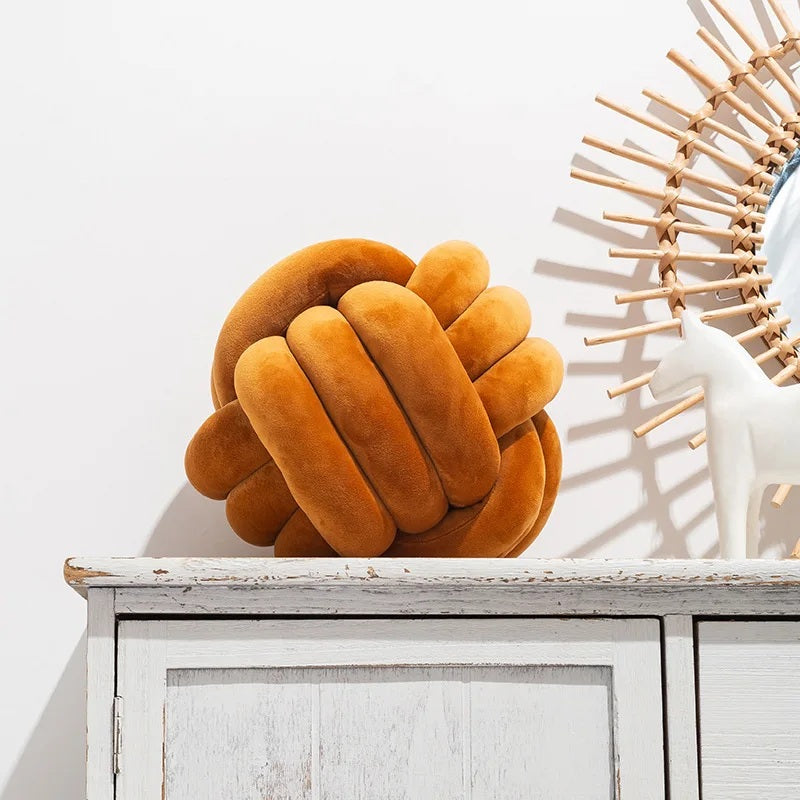 Coussin décoratif tressé pour lit de bébé en forme de nœud, de couleur caramel chaud, posé sur un tapis rayé noir et blanc, devant un berceau blanc dans une chambre d'enfant de style scandinave.