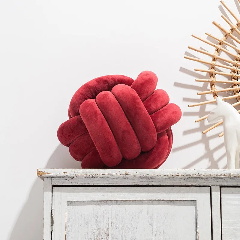 Coussin décoratif pour lit de bébé en forme de nœud tressé, de couleur rouge vin profond, posé sur un tapis rayé noir et blanc devant un berceau blanc dans une chambre d'enfant de style scandinave.
