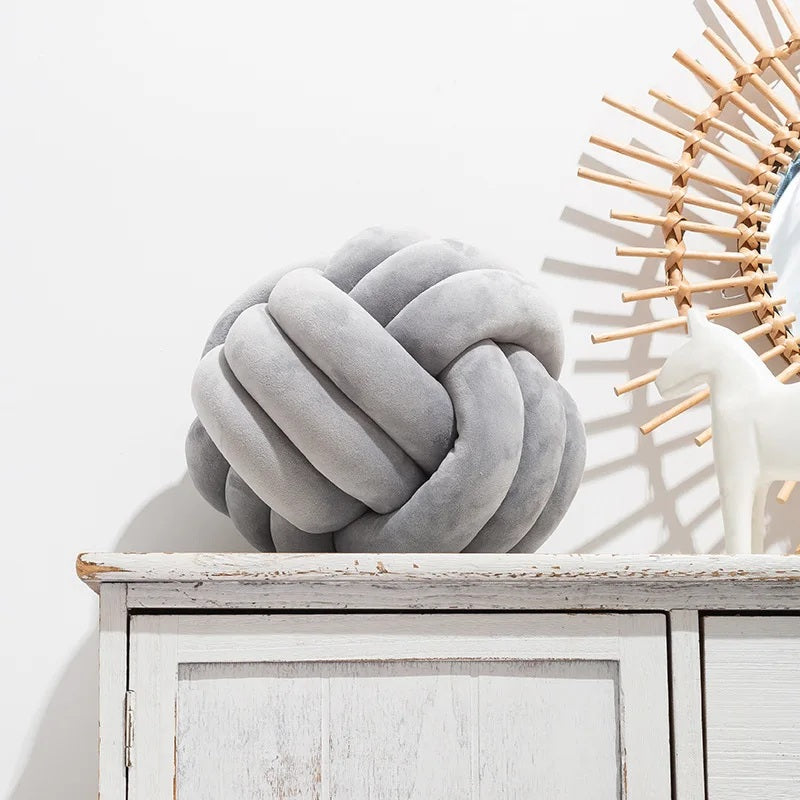 Une photo d'une chambre de bébé de style scandinave avec deux grands coussins en forme de nœud de couleur mauve poussiéreux, posés sur un tapis rayé noir et blanc à franges. Un berceau blanc avec une couverture tricotée se trouve en arrière-plan.