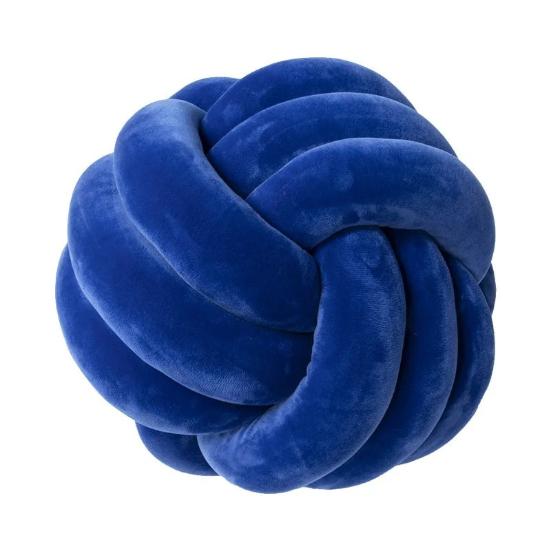 Coussin décoratif tressé en forme de nœud, de couleur bleu Klein vif, posé sur un tapis rayé noir et blanc, devant un berceau blanc et une chambre de bébé de style scandinave.