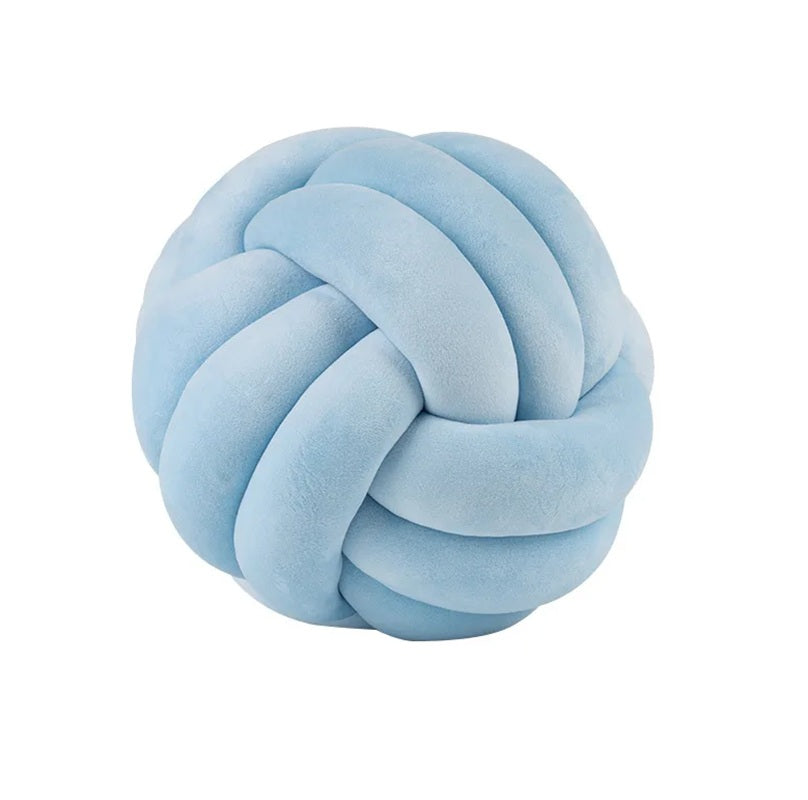 Coussin décoratif tressé en forme de nœud, de couleur bleu glacier doux, posé sur un tapis rayé noir et blanc devant un berceau blanc dans une chambre d'enfant de style scandinave.