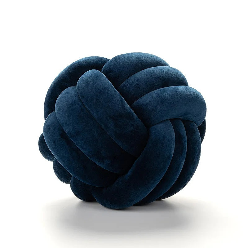 Coussin décoratif tressé en forme de nœud, de couleur bleu marine, posé sur un tapis rayé noir et blanc devant un berceau blanc dans une chambre d'enfant de style scandinave.