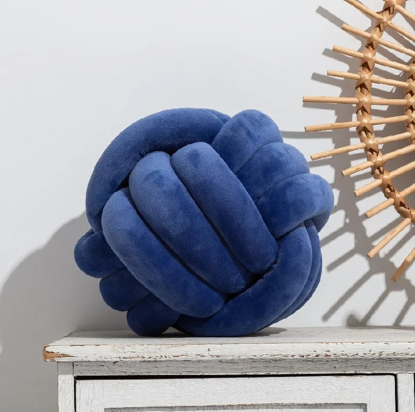 Coussin décoratif en forme de nœud tressé, de couleur bleu marine profond, posé sur un tapis rayé noir et blanc, devant un berceau blanc dans une chambre d'enfant de style scandinave.