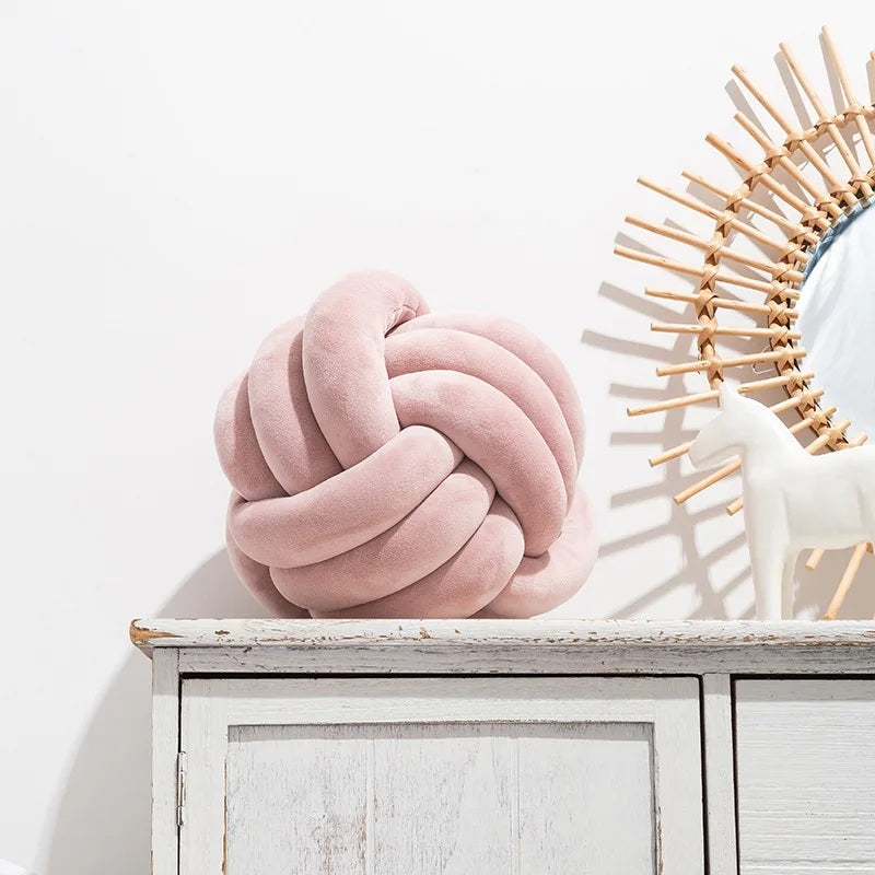 Coussin décoratif pour lit de bébé en forme de nœud, fabriqué en similicuir de couleur rose, posé sur un tapis rayé noir et blanc devant un berceau blanc dans une chambre d'enfant de style scandinave.