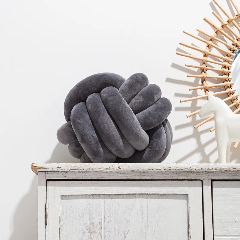 Un grand coussin décoratif pour lit de bébé en forme de nœud tressé, de couleur gris foncé, posé sur un tapis rayé noir et blanc devant un berceau blanc dans une chambre d'enfant de style scandinave.