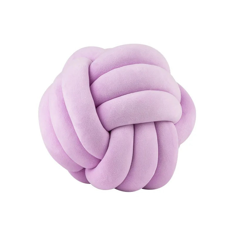Deux grands coussins décoratifs pour lit de bébé en forme de nœud, de couleur lilas, posés sur un tapis rayé noir et blanc devant une chambre d'enfant de style scandinave.