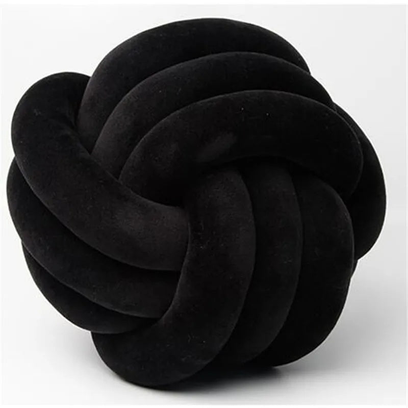 Coussin décoratif pour lit de bébé en forme de nœud, de couleur noire, fabriqué en tissu velours doux, posé sur un tapis rayé noir et blanc devant un berceau blanc dans une chambre d'enfant de style scandinave.