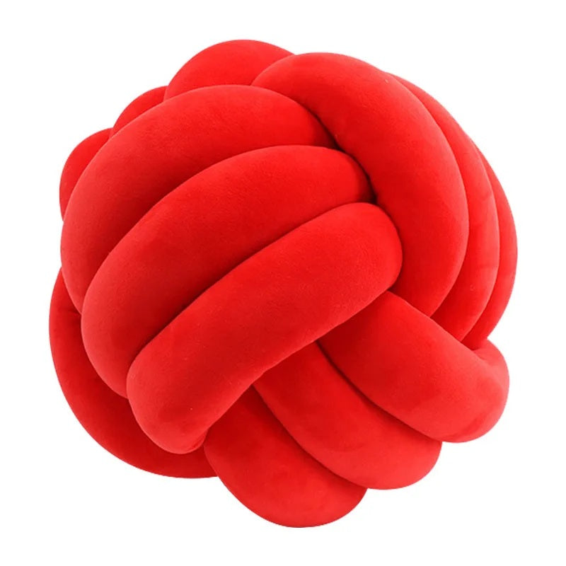 Un grand coussin décoratif pour lit de bébé en forme de nœud tressé, de couleur rouge vif, posé sur un tapis rayé noir et blanc, devant un berceau blanc et une chambre d'enfant de style scandinave.