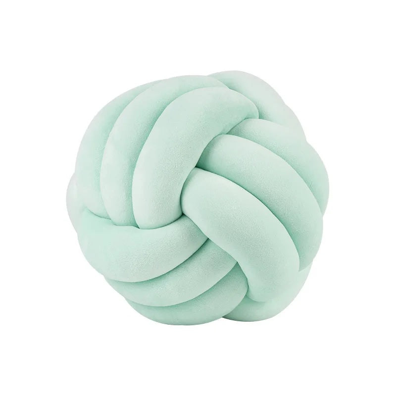 Coussin décoratif tressé en forme de nœud, de couleur vert clair doux, posé sur un tapis rayé noir et blanc devant un berceau blanc dans une chambre d'enfant de style scandinave.