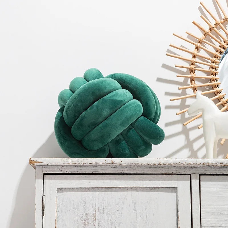 Coussin décoratif tressé en forme de nœud, de couleur vert sapin profond, posé sur un tapis rayé noir et blanc devant un berceau blanc dans une chambre d'enfant de style scandinave.
